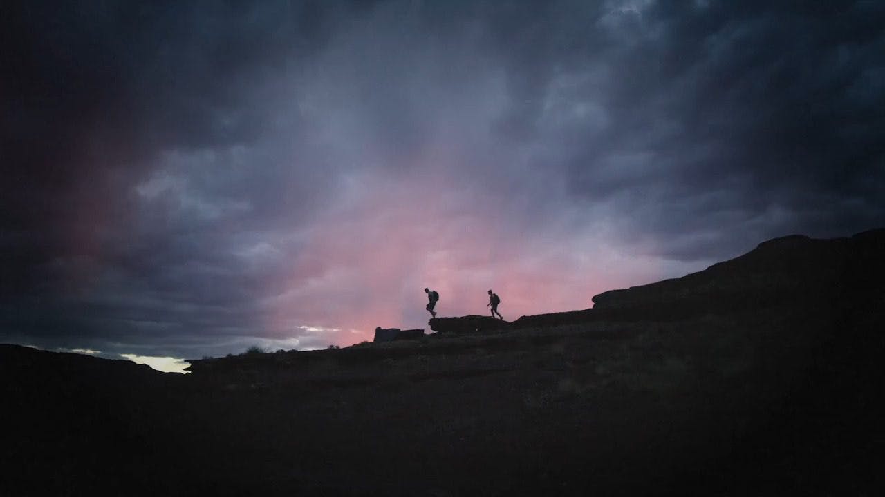 two people walking along the top of a hill with blue and pink skies