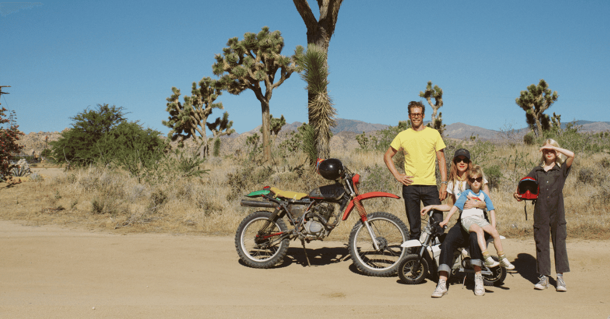 A man, woman, and two blonde little girls are posed in a group with a large dirtbike and a small dirtbike in the desert, there are trees in the background.