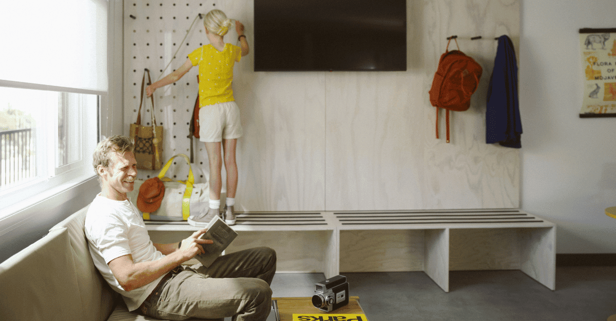 A young girl stands on a wooden bench next to a TV on the wall while a man smiles to someone else and is holding a book