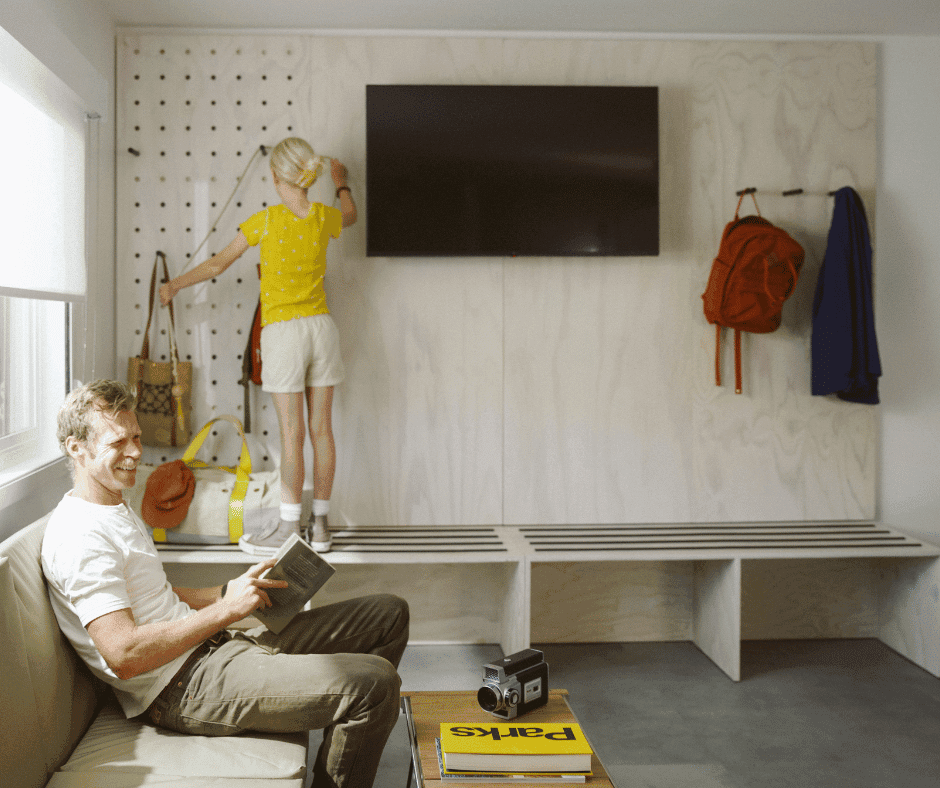 A young girl stands on a wooden bench next to a TV on the wall while a man smiles to someone else and is holding a book